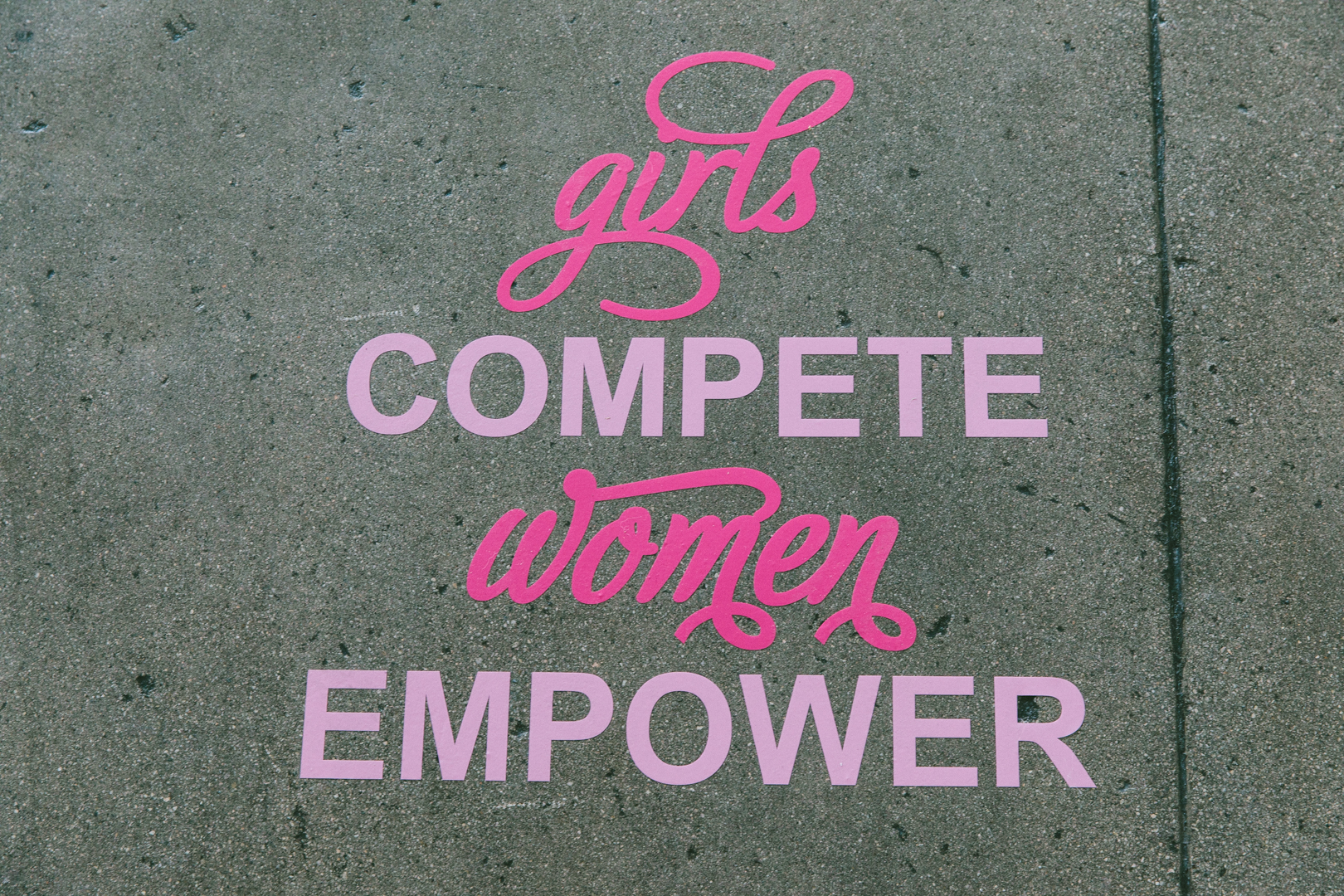 Sidewalk decal reading girls compete, women empower in pink script and lettering