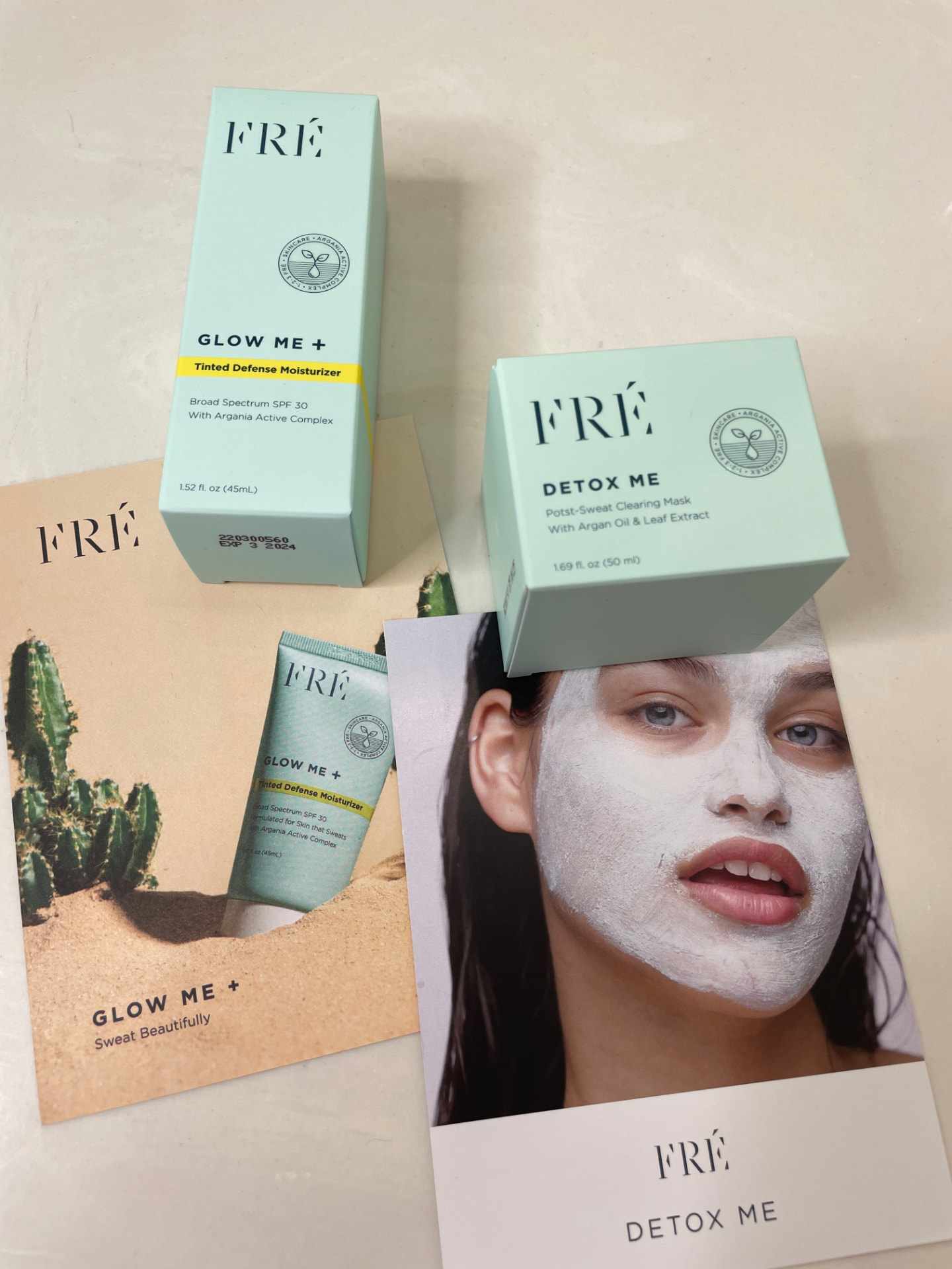 FRÉ Glow Me + tinted defense moisturizer and Detox Me clearing mask with brand cards on a marble counter