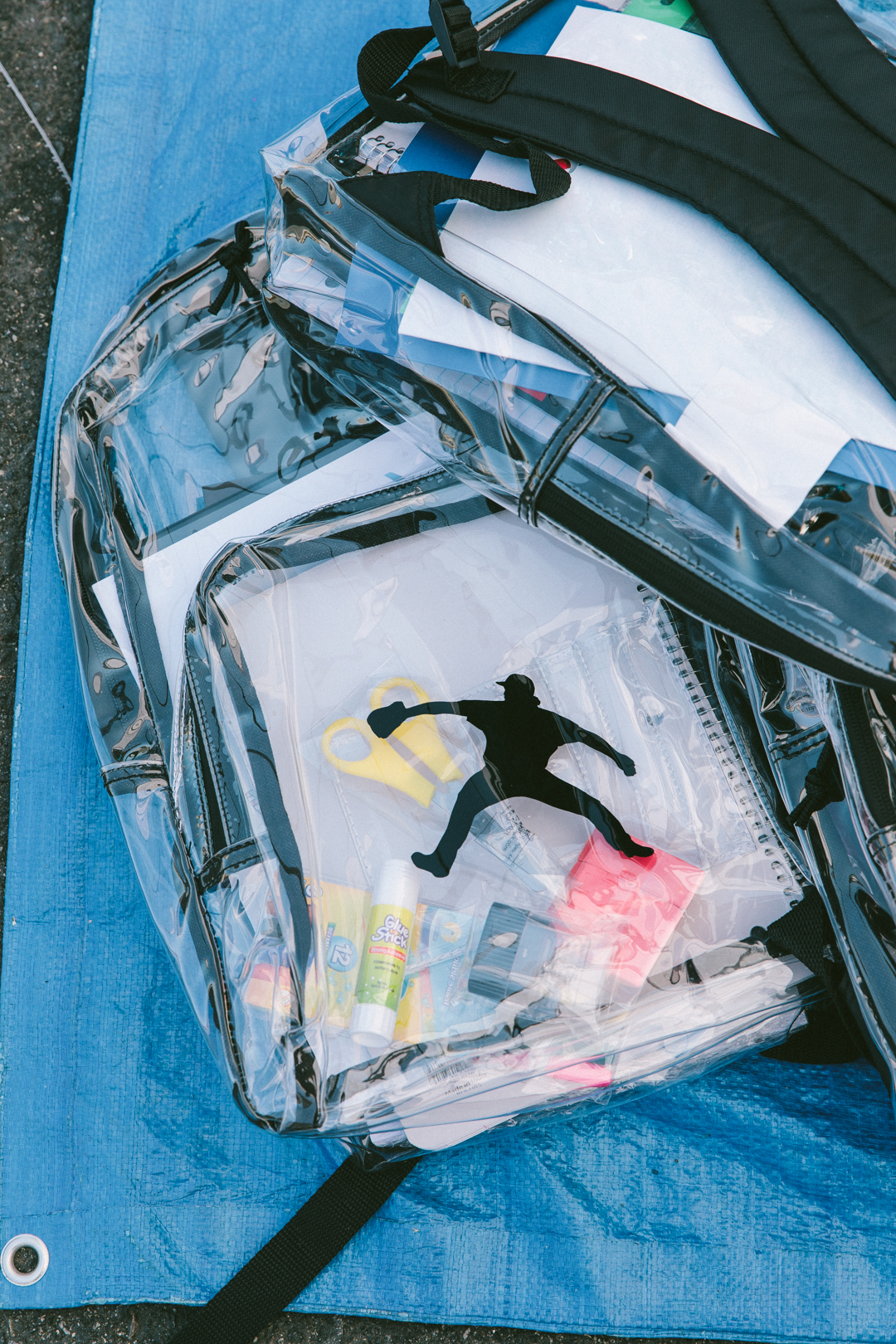 Clear backpack packed with notebooks, glue sticks, scissors and highlighters on a tarp