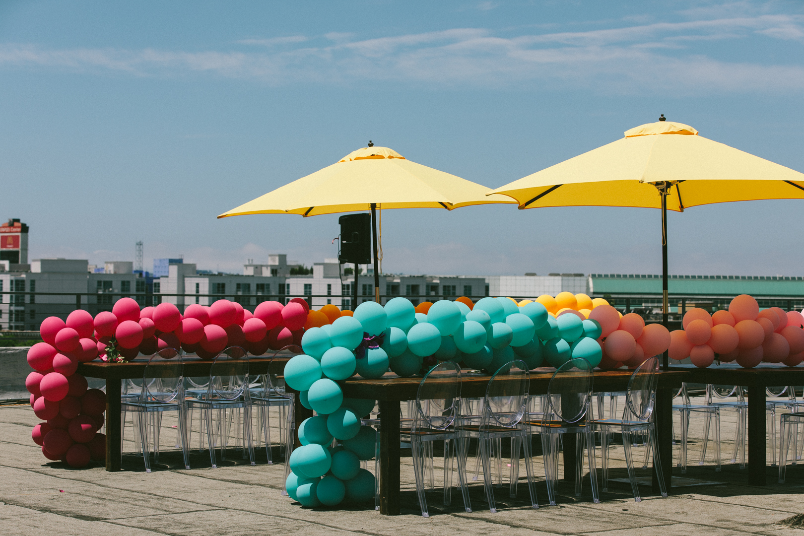 Rooftop dining tables under yellow umbrellas lined with balloon garlands and ghost chairs