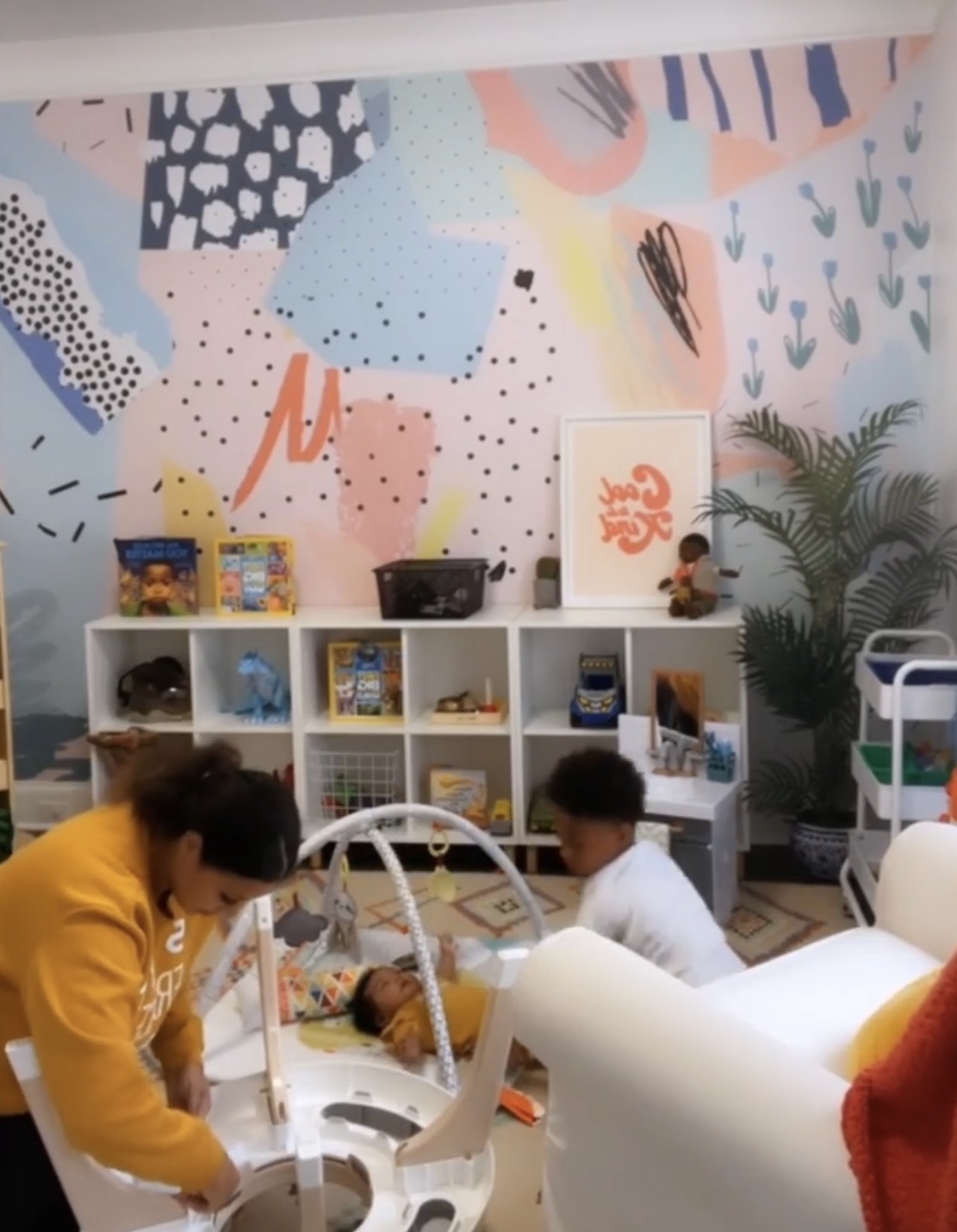 Playroom with a pastel mural, cube shelving of toys, a baby in a bouncer and an older sibling playing while LeLe Martin sets up